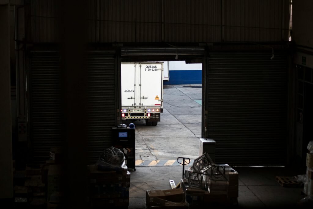 A white delivery truck exits a dimly lit warehouse loading dock, surrounded by packages and pallets.