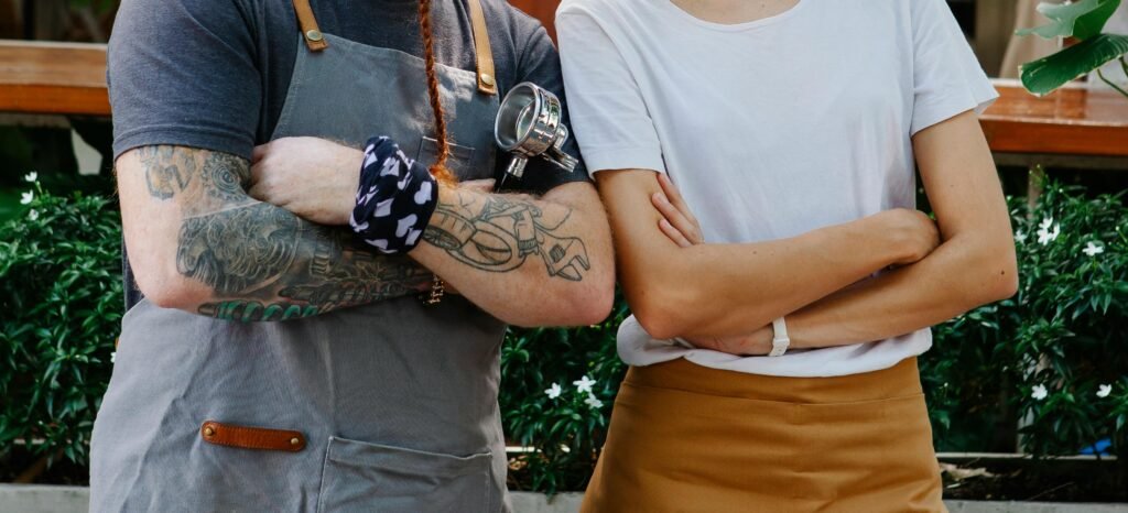 Crop anonymous couple of barista coworkers in aprons and casual clothes spending time in street with portafilter near plants in daytime