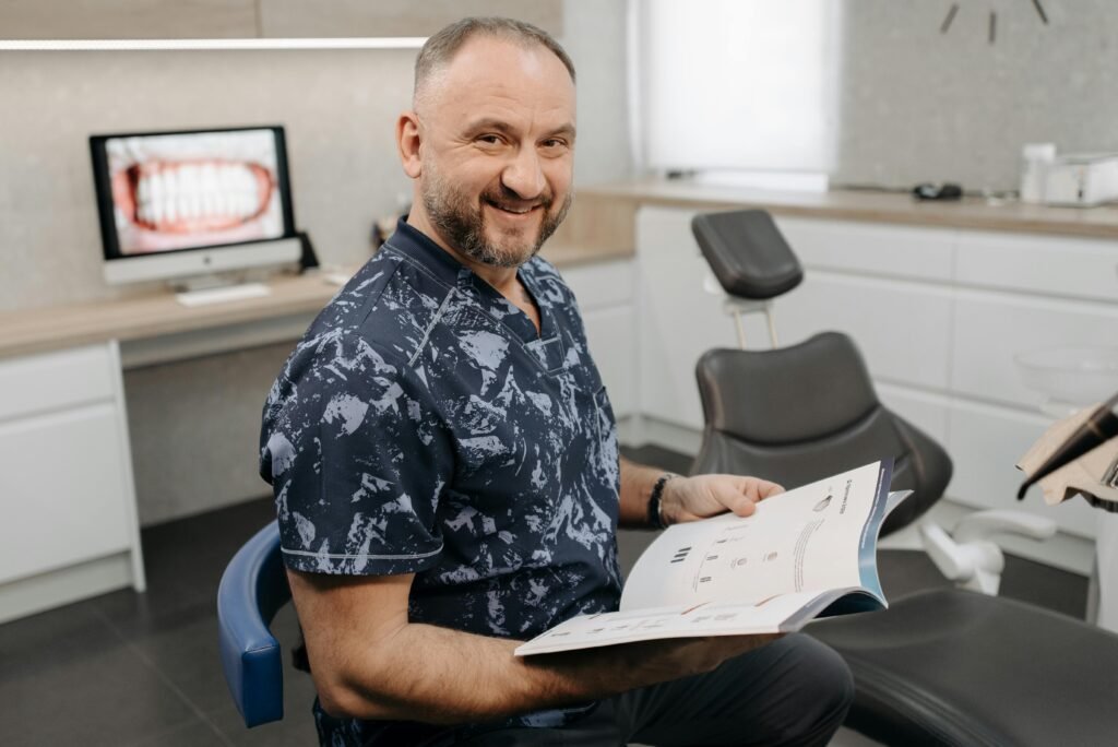 Smiling dentist holding a book, sitting in a dental office with equipment around.