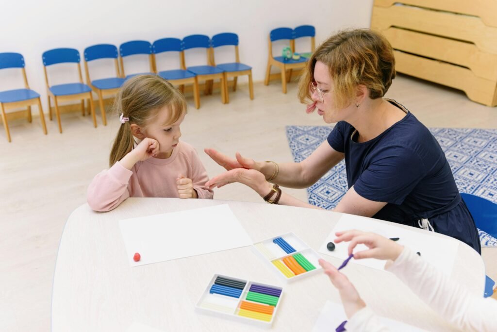 A teacher helps a child with a clay molding activity in a bright kindergarten classroom.